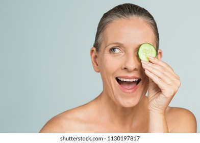 Portrait Of Mature Beautiful Naked Woman Smiling Hiding Eye Behind Cucumber Slice. Beauty Portrait Isolated On Gray Background. Funny Senior Woman In A Playful Mood Covering Eyes With Cucumber Slice.