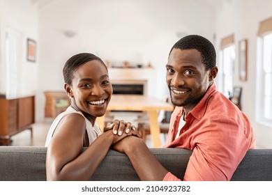 Portrait Of Mature Black Couple Looking At Camera While Sitting On Sofa. Happy African American Couple Relaxing At Home. Middle Aged Man Holding Hands Of His Beautiful Ethnic Wife While Smiling.