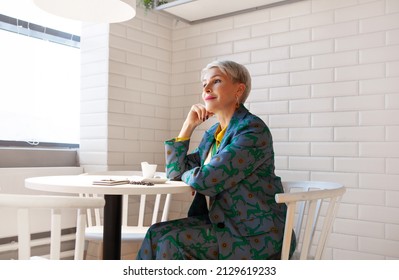 Portrait Mature Business Woman Thinking Or Dreaming In White Interior Cafe. Stylish Short Gray Hair Female In Blue Business Suit Sit At Table With Cup Of Coffee. White Brick Wall Background