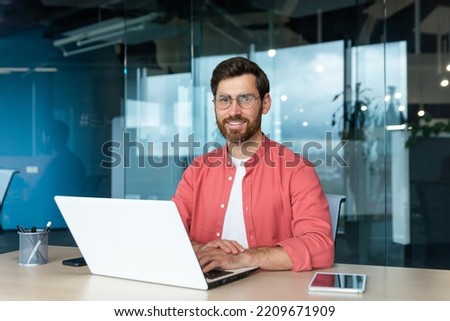 Portrait of mature businessman freelancer startup, bearded man smiling and looking at camera, business owner working inside modern office building wearing red shirt and glasses