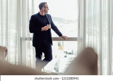 Portrait of mature businessman standing by a window and looking outside with a drink in hand. Man in business suit relaxing in hotel room with a drink in hand.