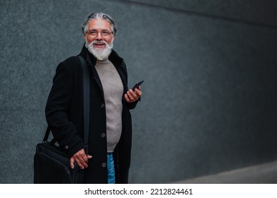 Portrait Of A Mature Businessman Walking Outside With A Smartphone In His Hands While Going To A Meeting. Senior Bearded Man Dressed In Stylish Black Coat With A Business Bag Looking Away. Copy Space.