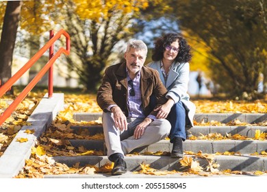 Portrait Of A Mature Couple 50 Years Old In An Autumn Park. Man And Woman Posing While Sitting On Concrete Steps 