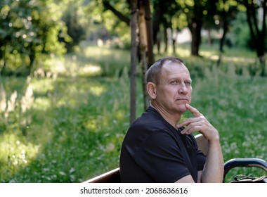 Portrait Of A Mature Courageous Man 60 Years Old, In A Black T-shirt, Sitting On A Bench, Against A Background Of Nature