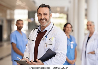 Portrait Of Mature Doctor Wearing Lab Coat And Stethoscope Reading Patient Case File In Hospital. Successful Head Physician Standing In Hospital Hallway And Looking At Camera With His Medical Team.