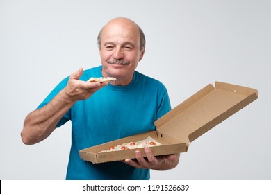 Portrait Of Mature European Man Eating A Slice Of Pizza. In His Hands He Holds A Box Of Food. Studio Shoot. Concept Of Eating Fast Food At Old Age