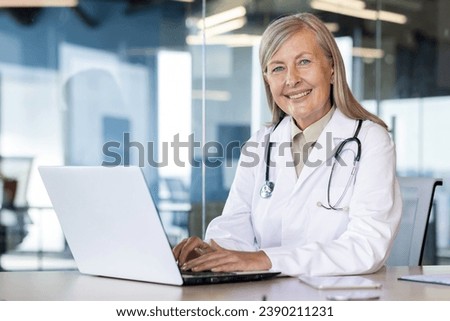 Portrait of mature experienced female doctor, gray-haired senior woman in white medical coat smiling looking at camera, consulting patients remotely, sitting at table with laptop inside clinic.