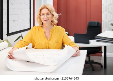 Portrait of mature female architect in eyeglasses looking at camera while working with blueprints of new buildings at table