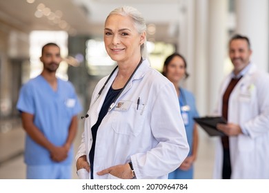 Portrait of mature female doctor on hospital corridor. Confident general practitioner standing in hospital hallway with her healthcare team in background. Successful head physician in private clinic. - Shutterstock ID 2070964784