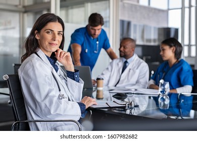 Portrait of mature female doctor sitting in meeting room with specialist and nurses discussing case in background. Successful woman doctor looking at camera with medical staff brainstorming.
