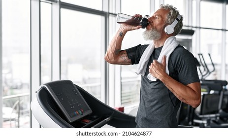 Portrait Of Mature Grey-haired Sportsman, Drinking Water From Bottle After Workout. Handsome Bearded Eldrely Man With White Towel On Shoulders Drinking To Stay Hydrated At Gym.