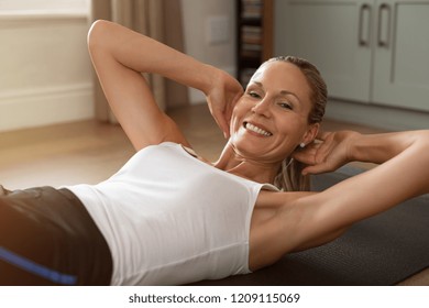 Portrait Of Mature Happy Blond Woman Doing Abdominal Exercises. Fitness Woman Doing Sit-ups At Home Lying On Yoga Mat. Blonde Lady Doing Body Exercises And Looking At Camera.