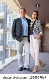 Portrait Of Mature Hispanic Businessman And Beautiful Hispanic Businesswoman Together By The Glass Window Of The Office Building Indoors