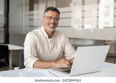 Portrait of mature Indian or Latin business man ceo trader using laptop computer, typing, working in modern office. Middle-age Hispanic smiling handsome businessman entrepreneur looking at camera. - Powered by Shutterstock