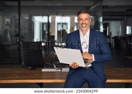 Portrait of mature Indian or Latin business man ceo trader using laptop computer, typing, working in modern office. Middle-age Hispanic smiling handsome businessman entrepreneur looking at camera.
