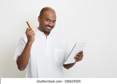 Portrait Of Mature Indian Man Shopping Online Using Tablet Pc, Making Payment By Credit Card. Asian Man Standing On Plain Background With Shadow And Copy Space. 