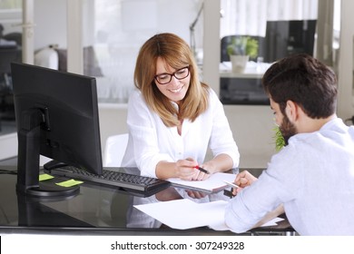 Portrait Of Mature Insurance Agent Giving Advise Her New Client While Sitting At Office In Front Of Computer. Businesswoman Making Financial Plan. 