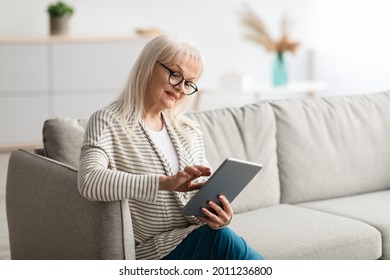 Portrait Of Mature Lady Wearing Glasses Spending Time With Digital Tablet At Home, Browsing Internet, Watching Videos, Checking Social Networks While Relaxing On Sofa In Living Room, Free Copy Space