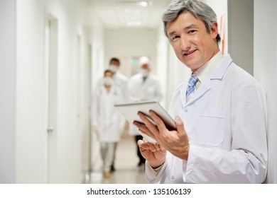 Portrait of mature male doctor holding digital tablet with colleagues walking in background