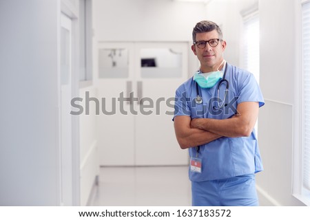 Portrait Of Mature Male Doctor Wearing Scrubs Standing In Hospital Corridor