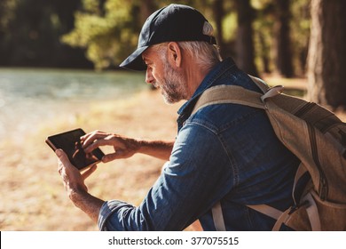 Portrait of mature male hiker using digital tablet outdoors. Senior man with backpack sitting by a lake looking at his digital tab for directions and location. - Powered by Shutterstock