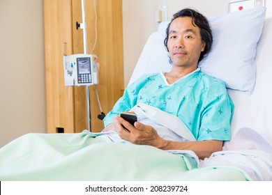 Portrait Of Mature Male Patient Holding Mobile Phone While Resting On Bed In Hospital