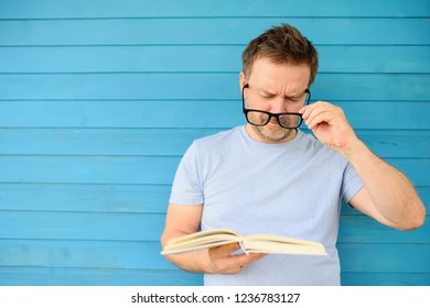 Portrait of mature man with big black eye glasses trying to read book but having difficulties seeing text because of vision problems. Problems disorder vision.