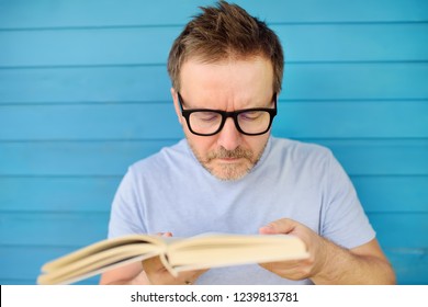 Portrait of mature man with big black eye glasses trying to read book but having difficulties seeing text because of vision problems. Problems disorder vision.