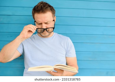 Portrait of mature man with big black eye glasses trying to read book but having difficulties seeing text because of vision problems. Problems disorder vision.