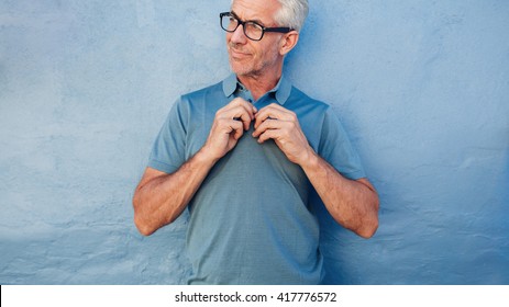 Portrait Of Mature Man Buttoning Up His Tshirt, He Is Standing Against A Wall And Looking Away.