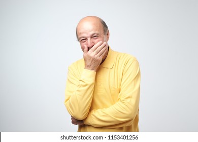 Portrait Of Mature Man Laughing And Covering His Mouth With Hand Over White Background. Trying To Hold A Laugh Not To Offend Some Person. He Just Heard A Funny Joke