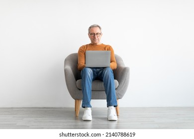 Portrait Of Mature Man Using Laptop Sitting On Armchair In Living Room. Confident Male Adult In Glasses Working On Pc, Typing On Keyboard Isolated On White Studio Wall, Full Body Length, Free Space