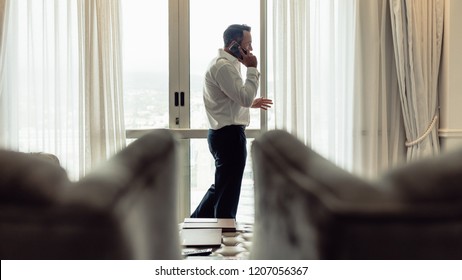 Portrait Of Mature Man Walking Inside A Hotel Room And Talking On Phone. Businessman Making Phonecall From Hotel Room On Business Trip.