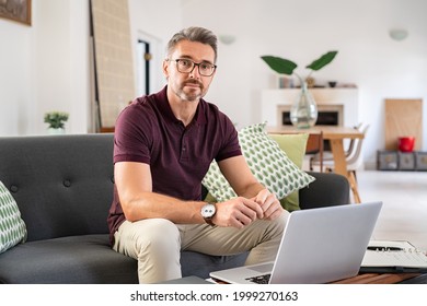 Portrait Of Mature Man Wearing Spectacles And Working From Home. Mid Adult Man Sitting On Sofa While Working On Laptop And Looking At Camera. Businessman Using Laptop In Telecommuting From Home.