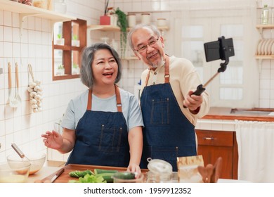 Portrait Of Mature Man And Woman Pensioner Using Smartphone Taking Photo Selfie Together In The Kitchen At Home. Cheerful Asian Senior Couple Having Fun Cooking Healthy Food Together. 