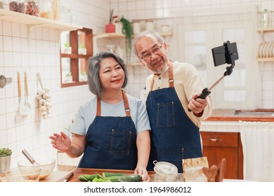 Portrait Of Mature Man And Woman Pensioner Using Smartphone Taking Photo Selfie Together In The Kitchen At Home. Cheerful Asian Senior Couple Having Fun Cooking Healthy Food Together. 