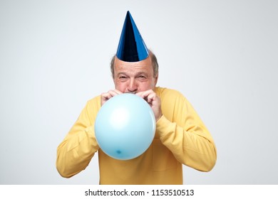 Portrait Of Mature Man In Yellow Tshirt And Funny Cap Blowing A Blue Balloon Over A White Background. Preparing For A Birthday Party