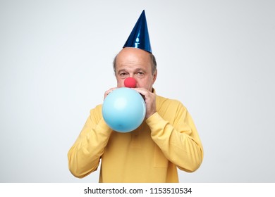 Portrait Of Mature Man In Yellow Tshirt And Funny Cap Blowing A Blue Balloon Over A White Background. Preparing For A Birthday Party