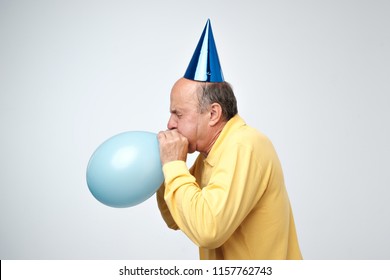 Portrait Of Mature Man In Yellow Tshirt And Funny Cap Blowing A Blue Balloon Over A White Background. Preparing For A Birthday Party. Side View