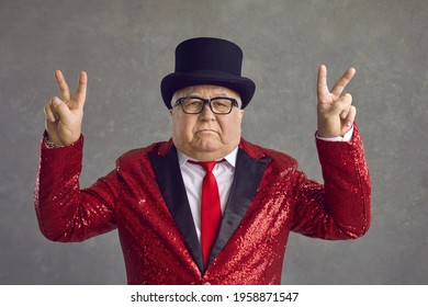 Portrait Of Mature Millionaire On Gray Background. Funny Rich Old Man In Spectacles, Bling Fancy Red Sequin Jacket And Black Top Hat Doing Peace Sign And Looking At Camera With Serious Face Expression