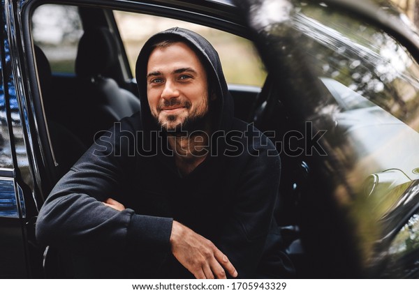 Portrait of\
mature neard man laughing and sitting in the car. Man taking a\
break on road trip. Man in black hoody look happy. Happy male\
driver smiling while sitting in a car.\
