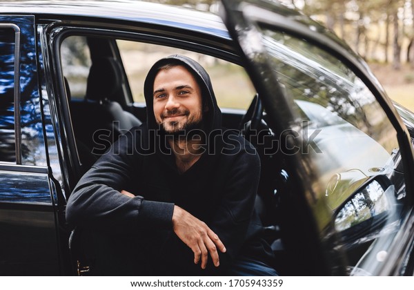 Portrait of\
mature neard man laughing and sitting in the car. Man taking a\
break on road trip. Man in black hoody look happy. Happy male\
driver smiling while sitting in a car.\
