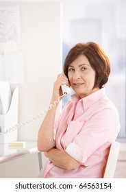 Portrait Of Mature Office Worker Talking On Land Line Phone At Desk, Looking At Camera.