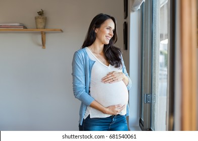 Portrait Of Mature Pregnant Woman At Home Looking Outside The Window. Young Pregnant Woman Standing Near Window At Home And Thinking About The Future. Smiling Pregnant Woman With Hands On Belly.