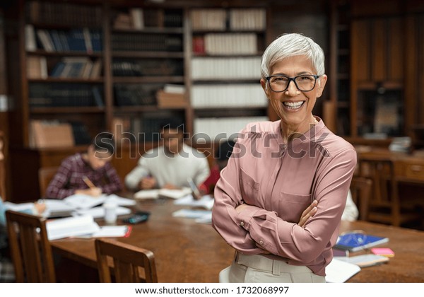 Portrait of mature professor standing in\
university library and looking at camera with copy space. Happy\
senior woman at the library working as a librarian. Satisfied\
college teacher\
smiling.
