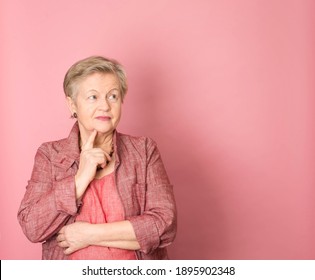 Portrait Of Mature Senior Woman Over Pink Background With Finger On Chin Thinking About Question, Pensive Expression. Smiling And Thoughtful Face. Doubt Concept.