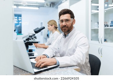 Portrait of mature successful scientist lab technician at workplace in laboratory with microscope, senior man with beard and goggles smiling and looking into camera, inventor with laptop . - Powered by Shutterstock