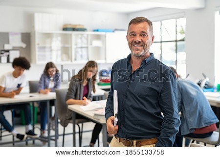 Portrait of mature teacher looking at camera with copy space. Happy mid adult lecturer at classroom standing after giving lecture. Satisfied high school teacher smiling while his students studying.