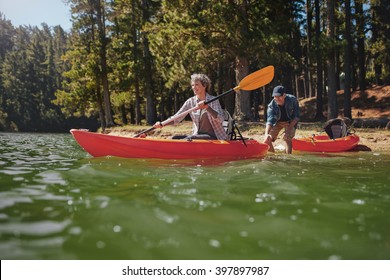 Portrait Of Mature Woman Canoeing In The Lake With Man About To Catch The Kayak From Behind. Senior Couple Having Fun Kayaking In The Lake.