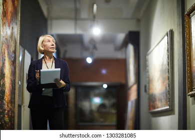 Portrait of mature woman holding clipboard looking at paintings standing in art gallery or museum, copy space - Shutterstock ID 1303281085